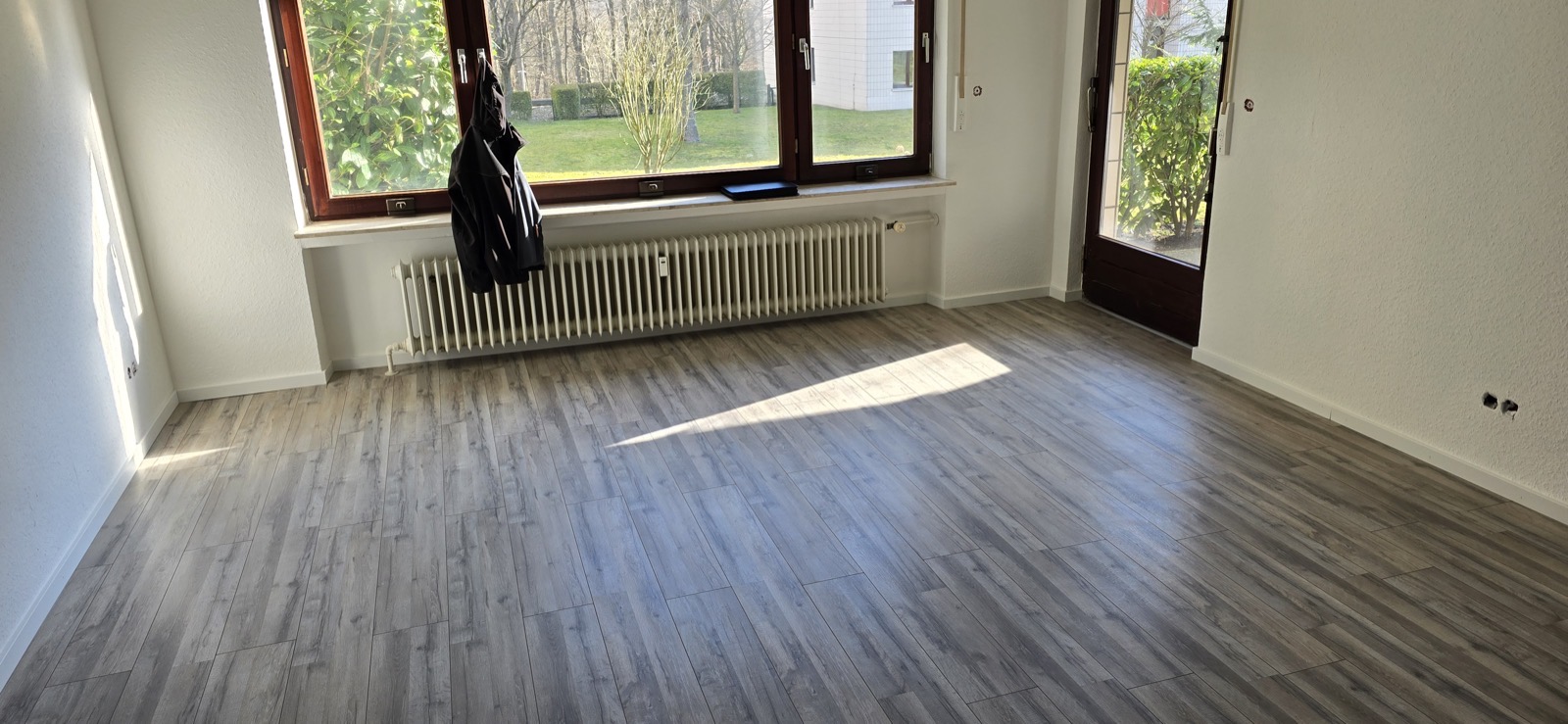 Fertig sanierter heller Wohnraum mit grauem Vinyl-Designboden, zwei Fenstern und Blick in den Garten — Hubertushöhe 8